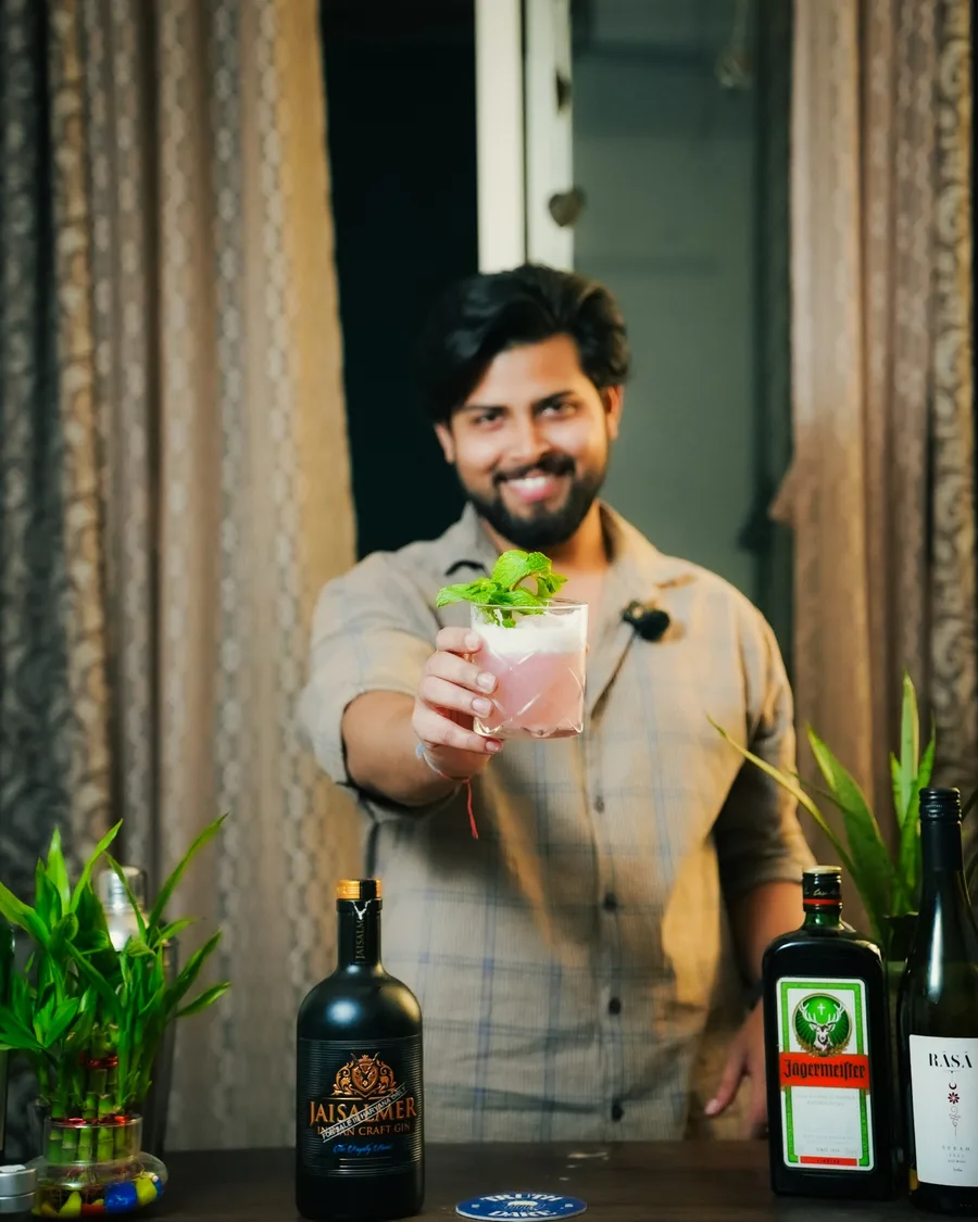 Yoginder smiling and holding out a pink cocktail garnished with mint, bottles on the bar in front of him